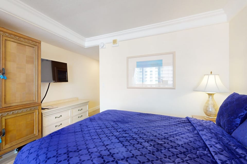 Relax in this inviting beach-stay bedroom featuring a plush bed with vibrant blue bedding, soft lighting, and resort-style comfort—perfect for unwinding after a day on the shore.