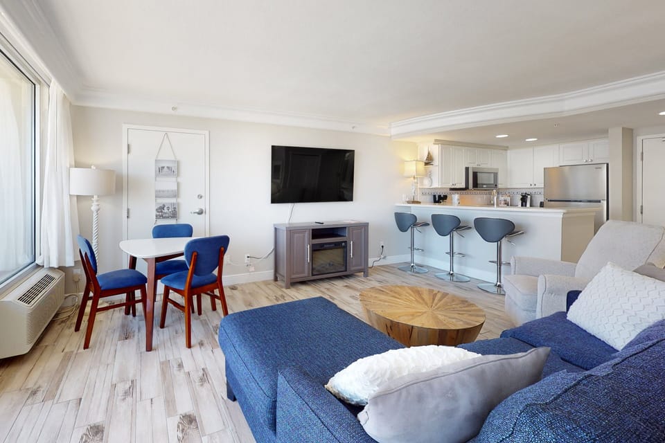 Relax in this airy beach-stay living space featuring a comfy sectional, dining nook, and open kitchen with bar seating. Large windows fill the room with coastal light—perfect for unwinding after a day by the ocean.