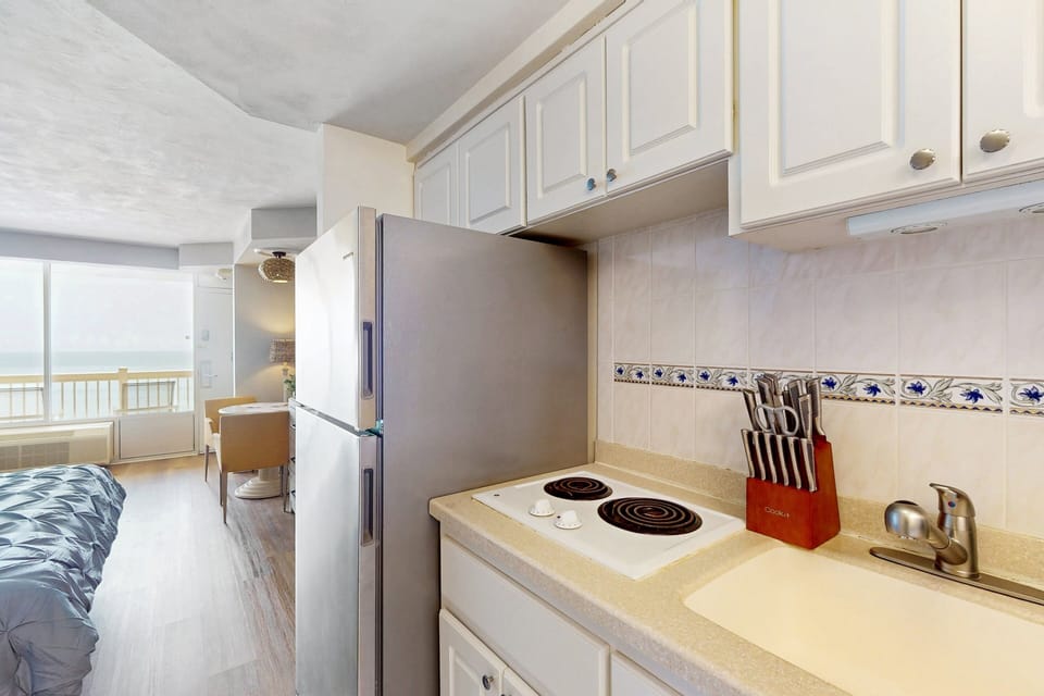 Compact beachside kitchenette featuring stovetop, fridge, and essentials. Enjoy convenient meal prep just steps from the sand, with a peek of the ocean view from your relaxing beach stay.