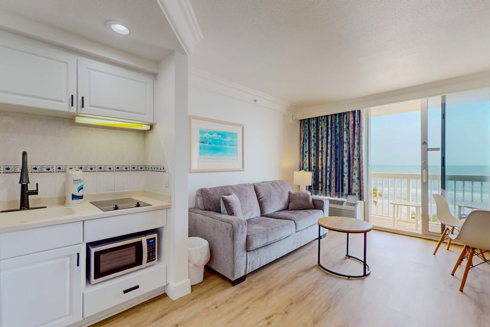 Relax in this bright beach-stay living area featuring a comfy sofa, convenient kitchenette, and sweeping ocean views from the balcony—perfect for unwinding after a day by the shore.