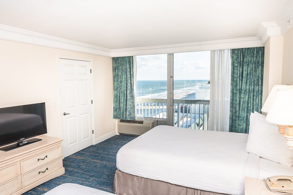 Bright oceanfront room featuring cozy beds, a flatscreen TV, and stunning beach views from large windows—perfect for relaxing after a sun-filled beach stay.