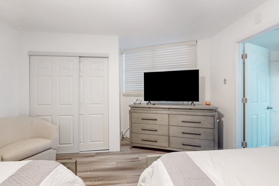 Relaxing coastal bedroom featuring cozy beds, dresser with TV, and ample storage. Ideal for unwinding during your beach stay after a full day of ocean, sun, and seaside fun.