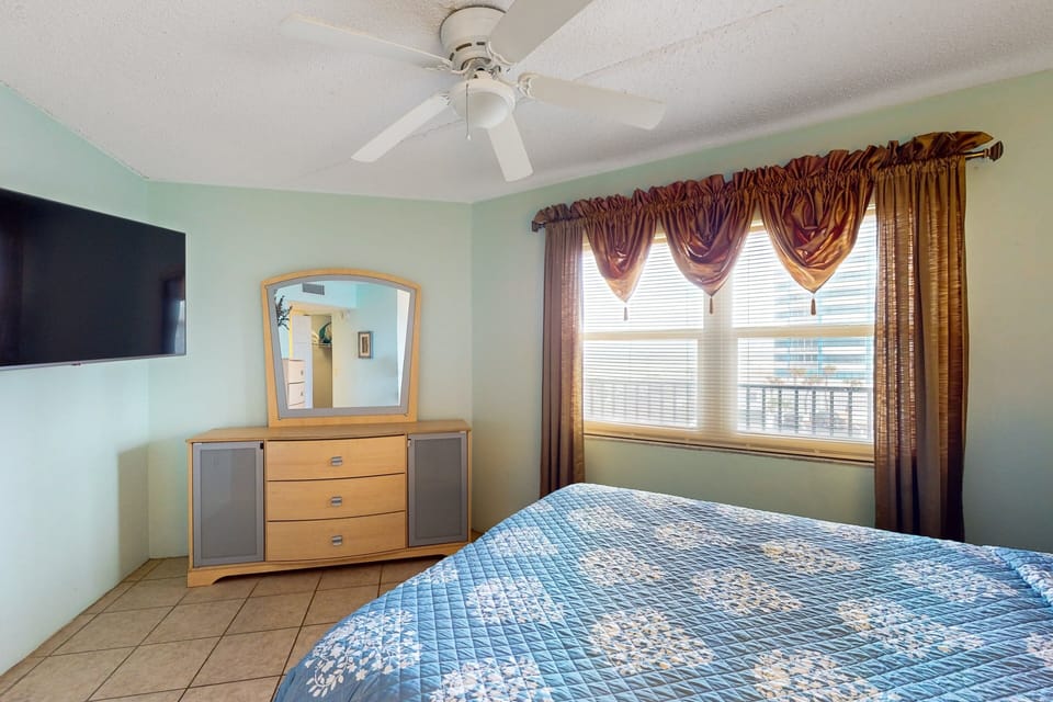 This relaxing beach stay bedroom features a cozy bed, ceiling fan, flat-screen TV, and soft coastal tones. A comfortable retreat to unwind and enjoy restful nights after your beach adventures.