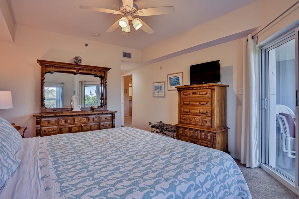 This relaxing coastal primary bedroom features a comfortable bed, TV, ample storage, and balcony access. Ideal for a peaceful beach stay, unwind in comfort after sunny days spent by the ocean.