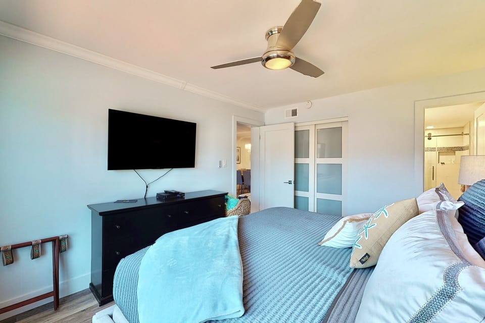 This comfortable coastal bedroom features a plush bed, ceiling fan, and flat-screen TV. A cozy retreat to unwind and enjoy quiet evenings during your relaxing beach stay.