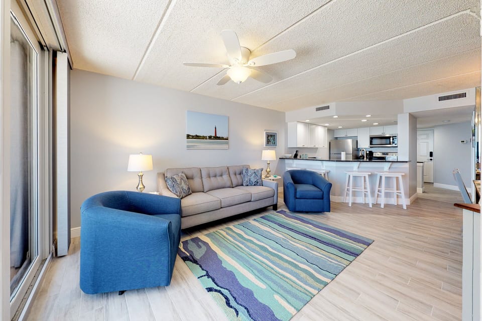This airy beach stay living room offers cozy seating, a vibrant coastal rug, and an open layout flowing into the kitchen—perfect for relaxing after a sunny day by the ocean.