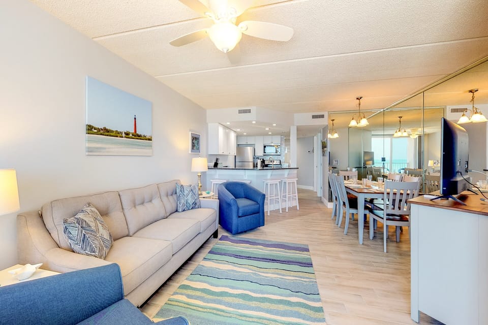 Enjoy this open beach stay layout featuring a comfy sofa, coastal accents, and a spacious dining area leading into a modern kitchen—perfect for relaxing and gathering after a day by the ocean.
