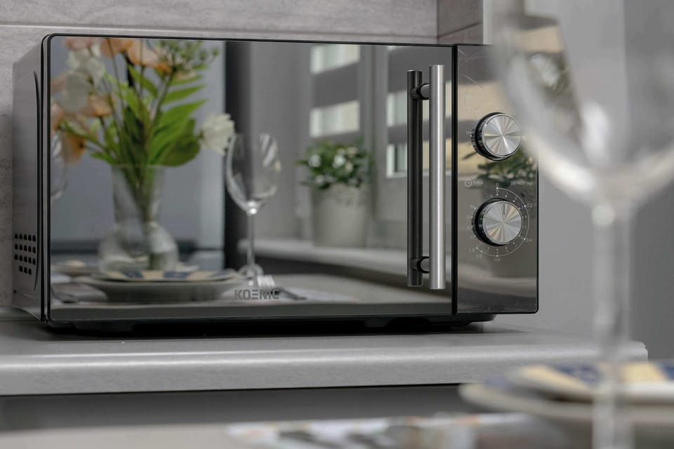 A macro shot of a microwave oven door reflecting glassware and greenery. The soft focus creates a modern and polished look. Shiny surfaces emphasize the cleanliness of the kitchen.