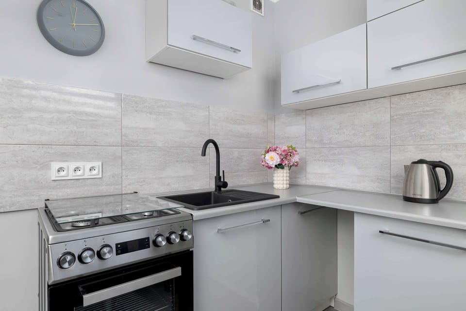 A close-up of a stove and countertop with a kettle and flower arrangement. Smooth white cabinets above provide storage. The light gray backsplash adds subtle texture.