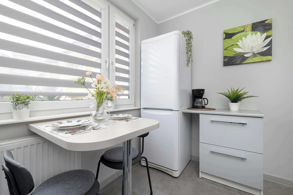 A dining nook with a white table, chairs, and decorative plants placed near the window. Roller blinds soften the incoming light. The minimalist setup feels peaceful and comfortable.