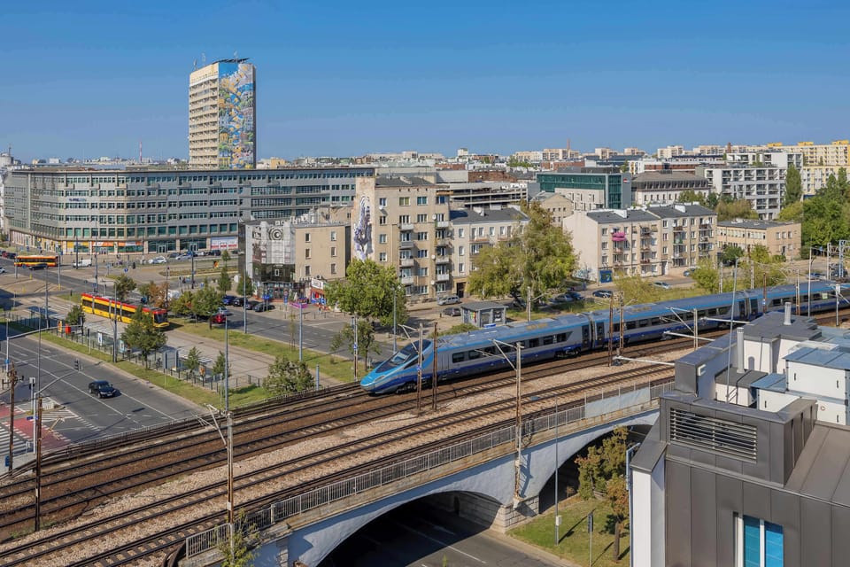 A high-level city view capturing railway tracks, apartment buildings, and greenery. The perspective gives a sense of urban movement and structure. The mix of modern buildings and natural elements creates visual harmony.