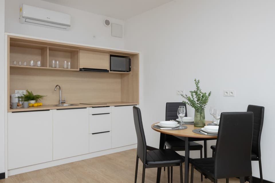 A small kitchen and dining area with white cabinets and wooden accents. A compact table with chairs offers space for meals or work. Soft lighting enhances the minimalistic design.