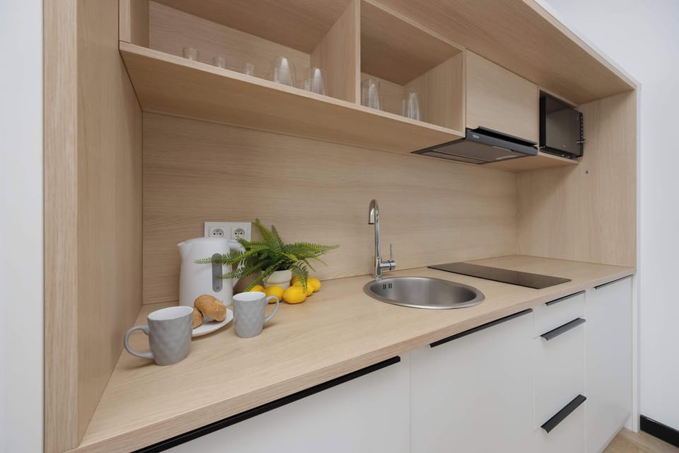 A functional kitchen counter equipped with a sink, stove, and light wooden shelving. Everyday essentials like cups and a cutting board are neatly arranged. The bright design creates a clean and practical food-preparation area.
