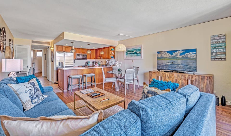 Open-concept living area with cozy blue sofas, rattan accents, and a dining space, seamlessly connected to a fully equipped kitchen with wooden cabinetry and modern appliances.