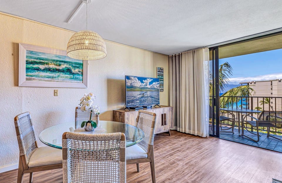 Dine in a bright and airy setting with a glass-top table, woven chairs, and soothing artwork, complemented by natural light streaming through sliding doors to the balcony.