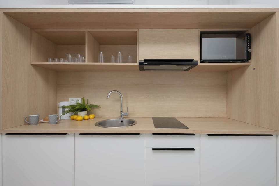 A modern kitchenette featuring wooden shelves, white cabinets, and neatly arranged appliances. A microwave and dishes are stored efficiently above the counter. The bright lighting keeps the space welcoming.