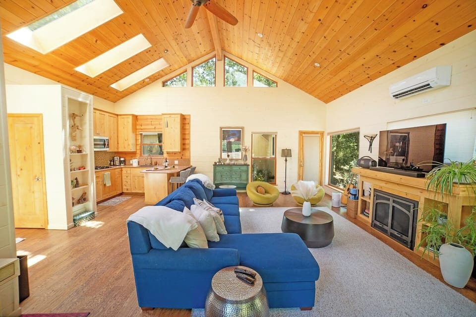 Dramatic vaulted wood ceiling with skylights floods this open-plan living space with golden light. Fireplace, blue sectional & seamless flow to the kitchen — relaxed luxury at its finest.