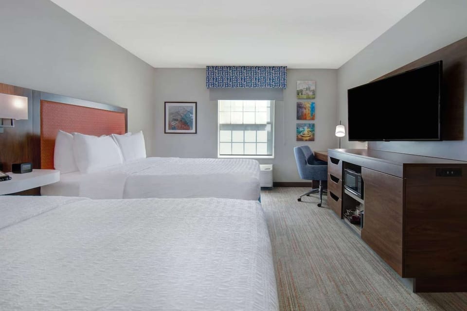 Bright room with two beds, desk, big TV, and wall art by the window