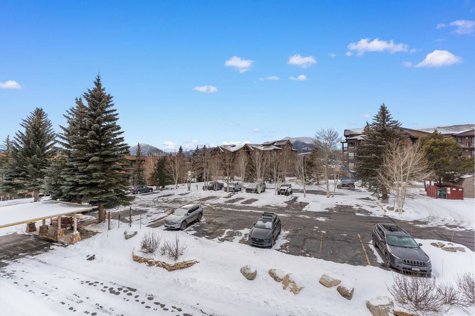 Snow-covered mountain resort parking area with evergreen trees and ski lodge buildings in winter setting.