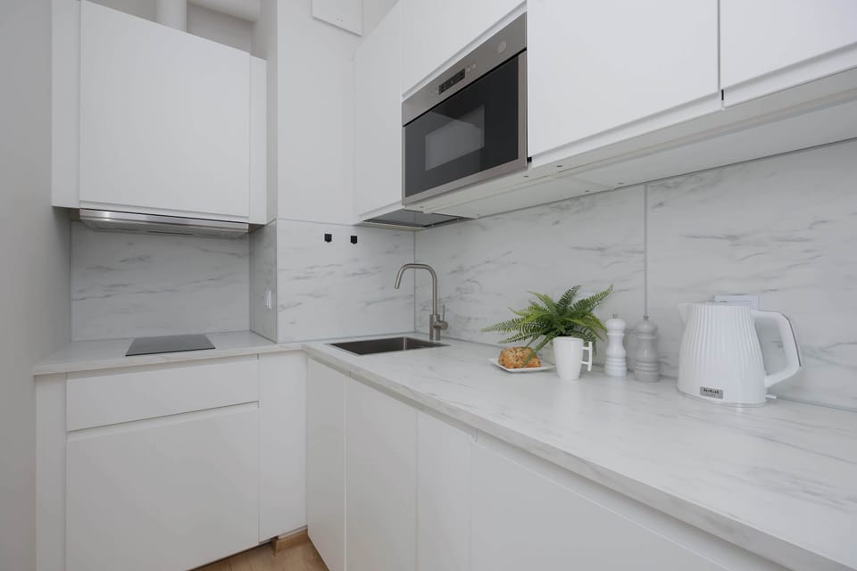 A modern white kitchen with sleek cabinets and built-in appliances. A small plant and simple accessories add a touch of freshness. The countertop layout emphasizes clean lines and functionality.