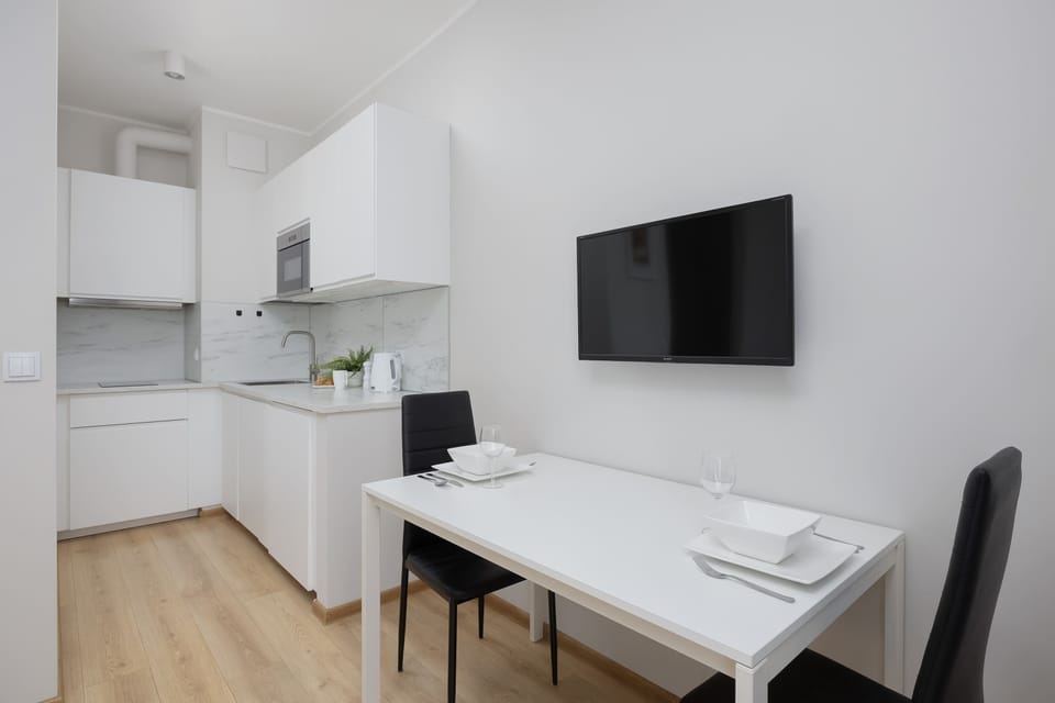 A compact kitchen and dining area with a white table and wall-mounted TV. Cabinets and appliances are neatly integrated into the wall. The layout looks efficient and space-saving.