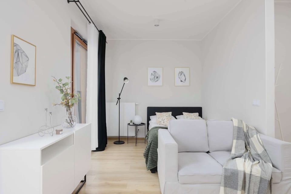 A living room angle featuring a white sofa, floor lamp, and bright natural light from the window. The room feels spacious and uncluttered. Light wood flooring adds warmth to the modern interior.
