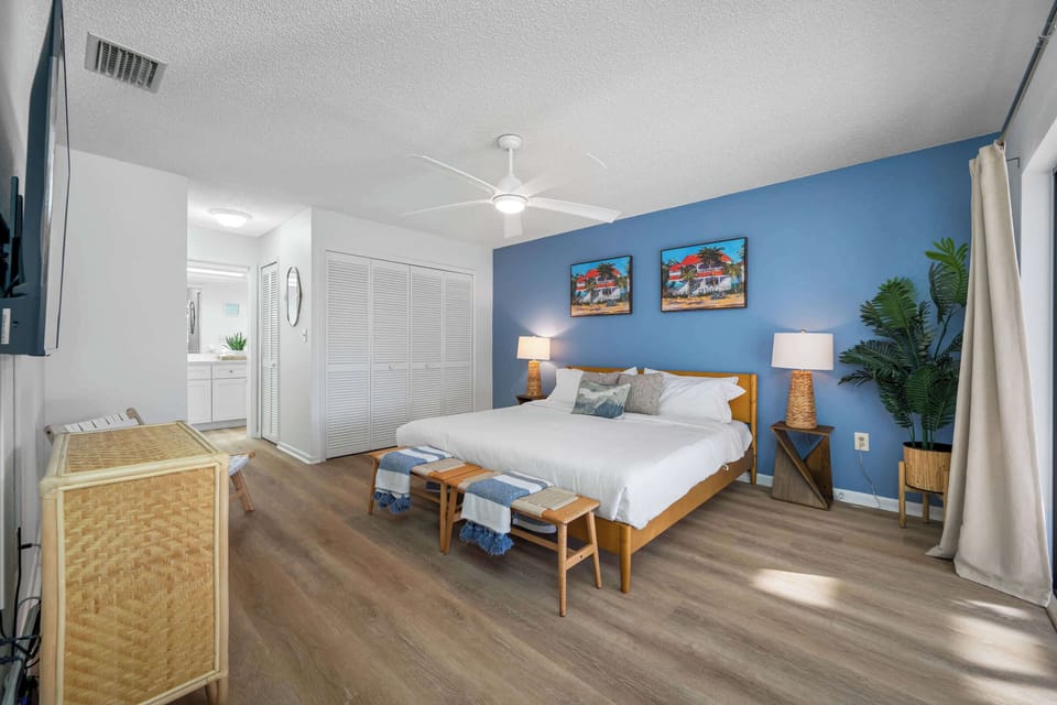 A blue accent wall and Caribbean streetscape prints give this king primary bedroom a bright, arty feel, ensuite just beyond. - Primary Bedroom - The Hammock - Largo, FL