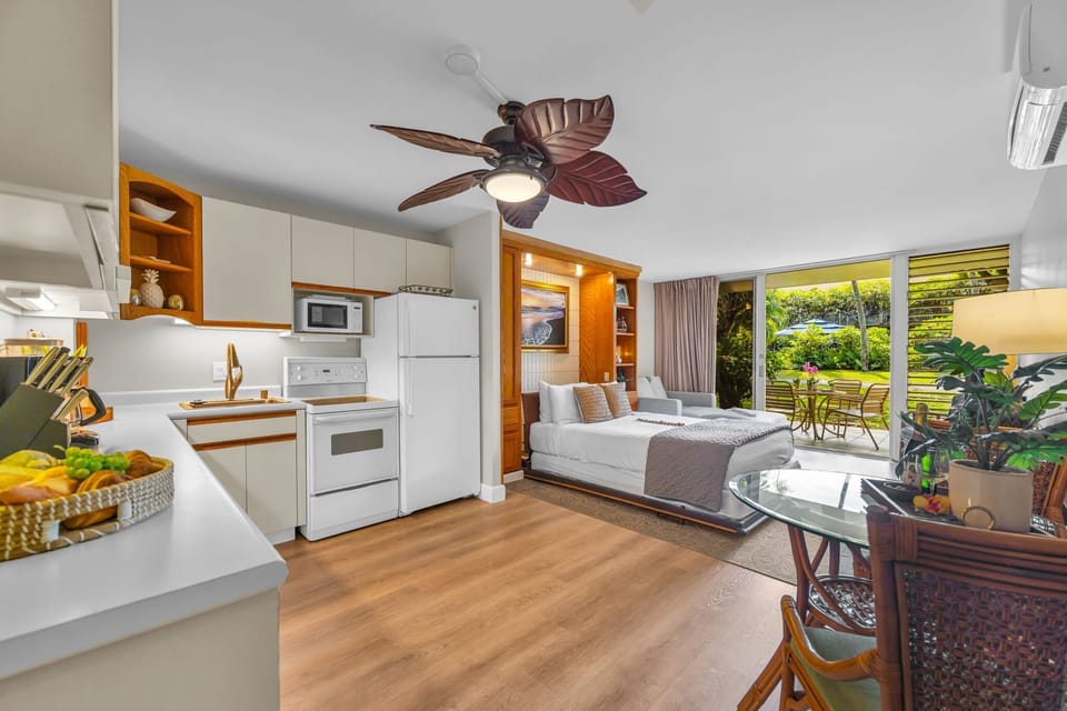 Relax in a bright studio featuring a cozy bed, well-equipped kitchen, and ceiling fan, complemented by natural light and garden views.
