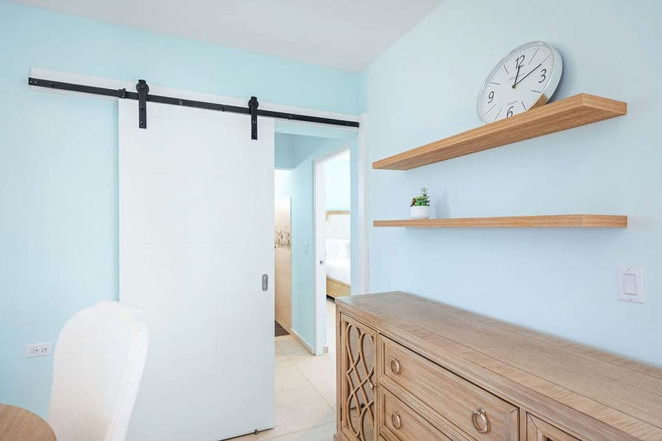 Just off the main dining area, a chic white barn door leads to the bedroom, while a rustic cabinet, floating shelves, and a large clock on soft blue walls combine style and function.