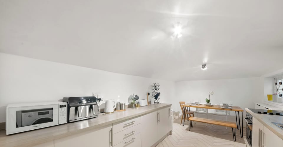 A bright, modern kitchen and dining area featuring light cabinetry and herringbone floors. A wooden dining set is tucked neatly under the sloped ceiling in the background.