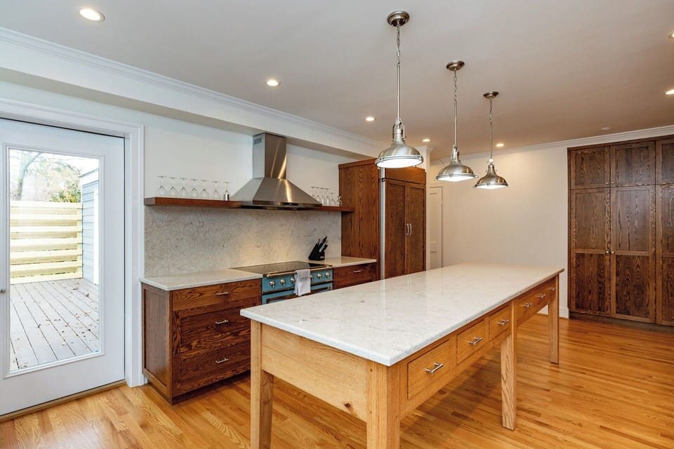 This modern kitchen showcases a large wooden island with white stone countertops and elegant pendant lighting. Professional stainless steel appliances and custom wood cabinetry create a high end culinary space.