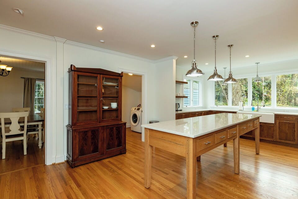 This gourmet kitchen features a stunning oversized wood island and bright white countertops. Stainless steel pendant lights hang over the workspace, while a classic dark wood hutch adds character and storage near the adjacent dining room.