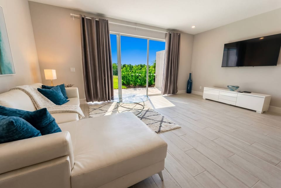 Bathe in natural light as you relax on the sleek white sofa, accented by plush teal pillows. The contemporary decor is enhanced by the expansive glass sliding doors, offering a glimpse of the lush greenery outside.