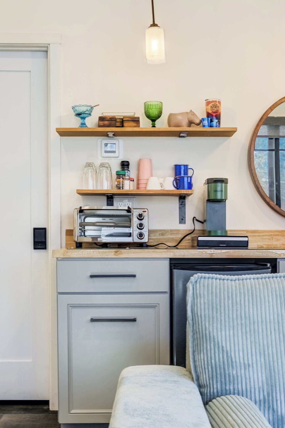 This view highlights the floating shelves with cups, glasses, spices, and decor above the toaster oven and Keurig, with cabinetry and the compact fridge below.