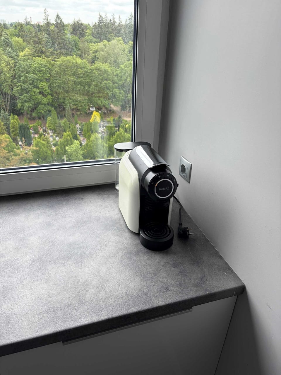 A compact coffee machine is placed on a gray windowsill next to a white wall. Large windows overlook a green, wooded landscape, giving the scene a calm and modern feel.