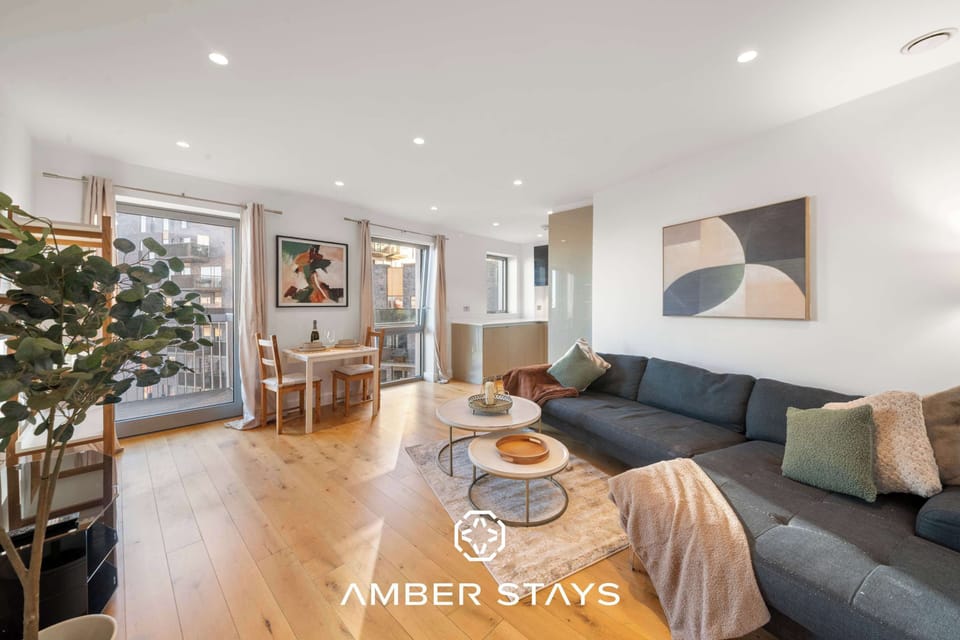 A bright, modern living room with light wood floors and white walls, featuring a large navy sectional and contemporary art. Floor-to-ceiling windows and recessed lighting create an airy, well-lit space.