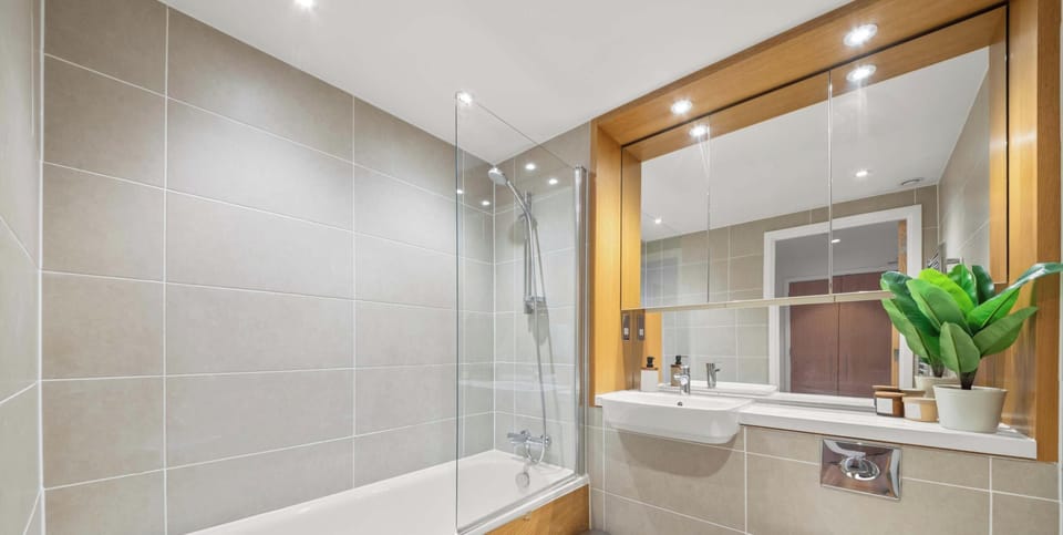 A modern, neutral bathroom featuring beige tiling, warm wood accents, and a sleek glass-enclosed tub. The compact space is efficiently designed with a wall-mounted toilet and a prominent chrome towel radiator.
