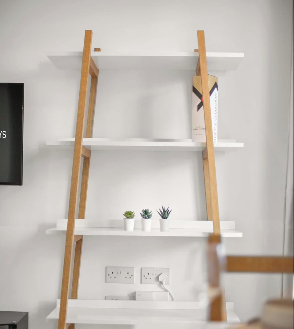 A minimalist, four-tier ladder bookshelf featuring light wood supports and white shelves. It is simply decorated with small plants and objects against a neutral wall.
