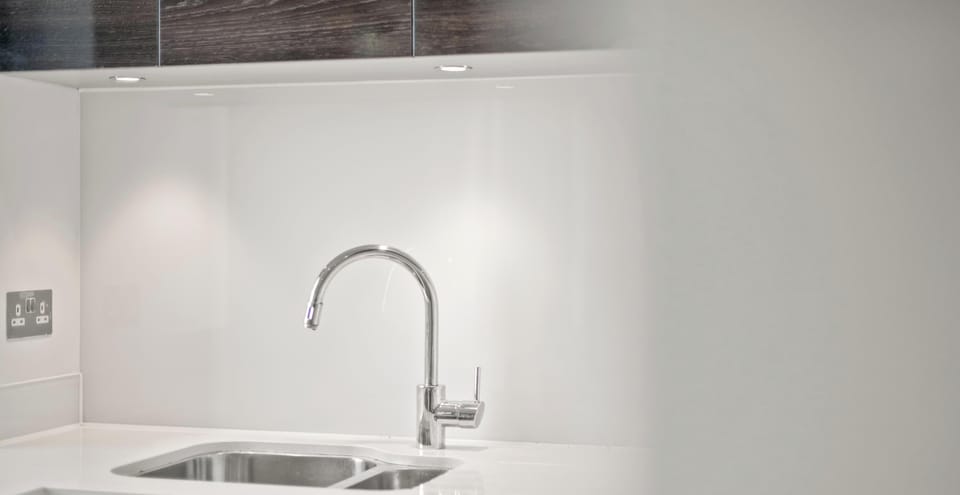 A modern, minimalist kitchen featuring a sleek chrome faucet and stainless steel sink. Bright under-cabinet lighting highlights the clean white countertops and backsplash.