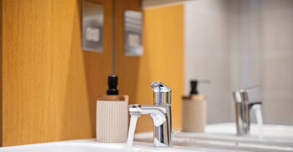 A sleek chrome faucet runs into a modern sink, accompanied by a ribbed beige soap dispenser. The clean setup is reflected in a large mirror against warm wood paneling.
