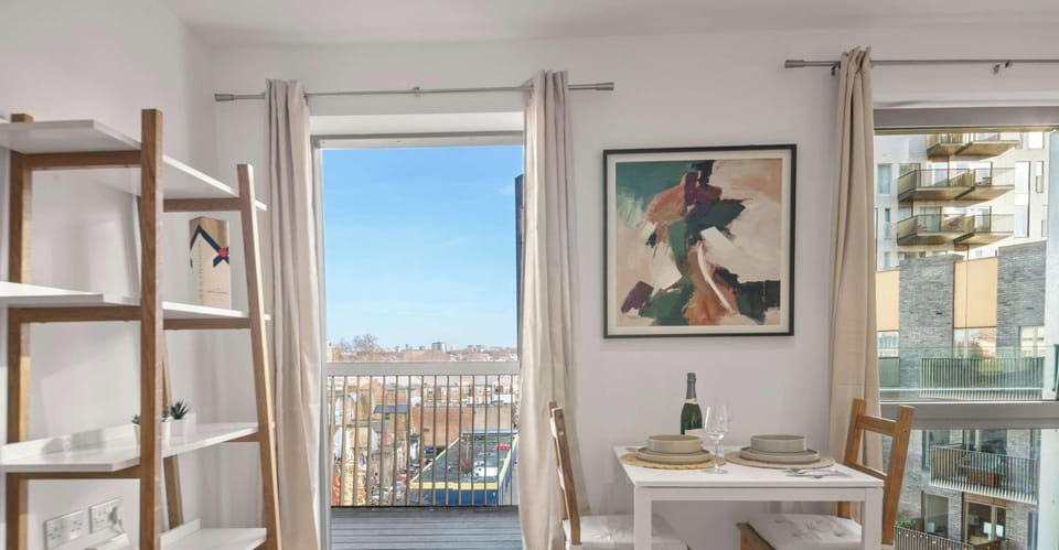 This bright and modern dining area features a white table set for two, framed by light wood chairs and a large abstract painting. A tall, open wooden bookshelf stands to the left, while large glass doors lead to a balcony with an expansive city view.