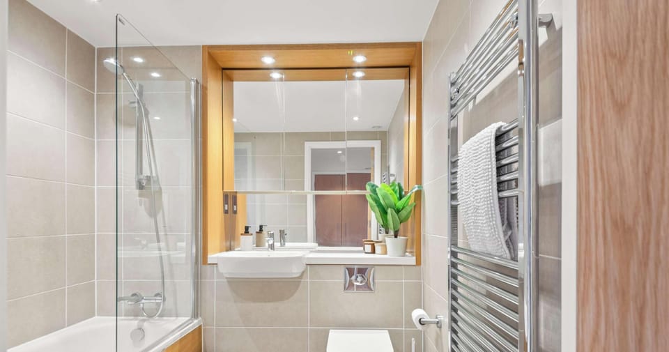 A modern, neutral bathroom featuring beige tiling, warm wood accents, and a sleek glass-enclosed tub. The compact space is efficiently designed with a wall-mounted toilet and a prominent chrome towel radiator.
