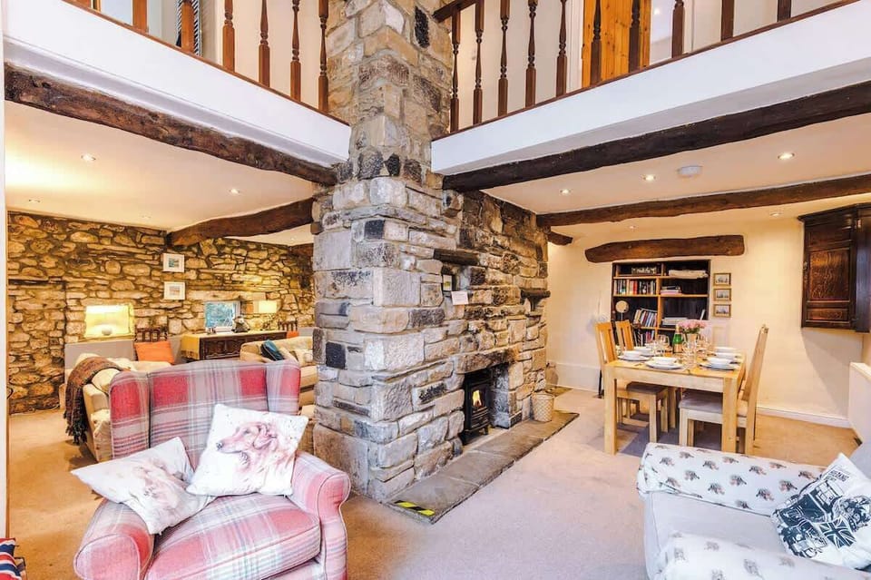 This open-plan living and dining area boasts a dramatic double-height ceiling anchored by a central stone fireplace and a rustic wooden mezzanine.