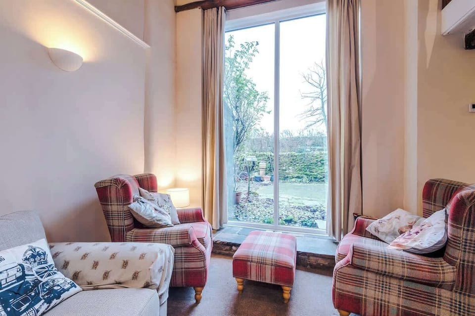 A sun-drenched seating nook features plaid armchairs and a matching ottoman arranged before a tall floor-to-ceiling window overlooking a garden.