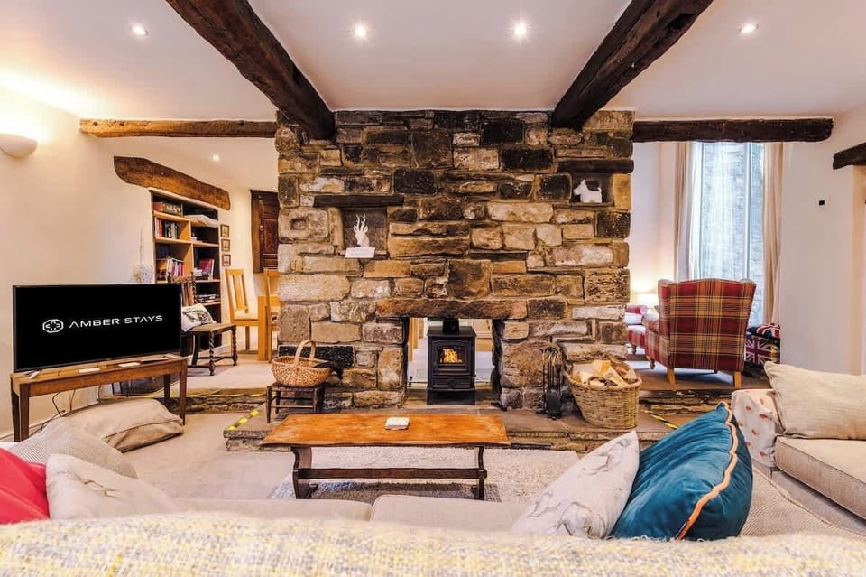 This cozy living space centers around a massive double-sided stone fireplace with a wood-burning stove, framed by rustic timber beams and comfortable, eclectic seating.