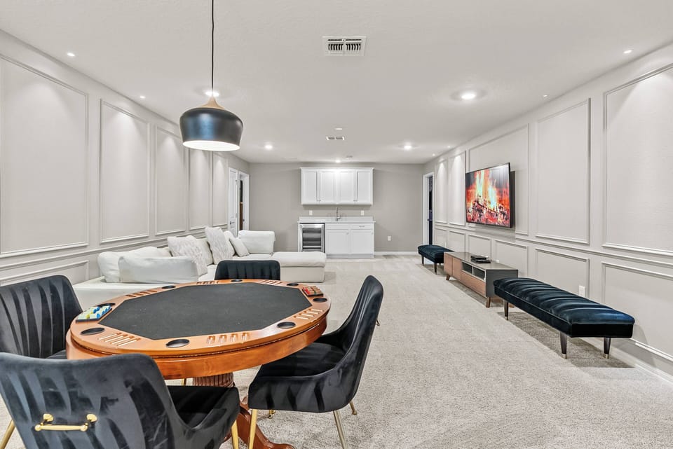 Enjoy a stylish living area with a sleek poker table and plush black chairs, ideal for gatherings or game nights. The soft gray carpet and illuminated paneling provide a cozy yet modern vibe, while the large TV adds to the fun.
