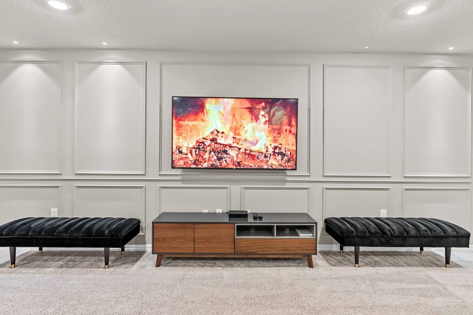 Relax in this stylish living room featuring plush black benches and a warm wooden media console on crisp white walls. The cozy fire imagery around the large TV creates an inviting atmosphere for unwinding after your adventures.