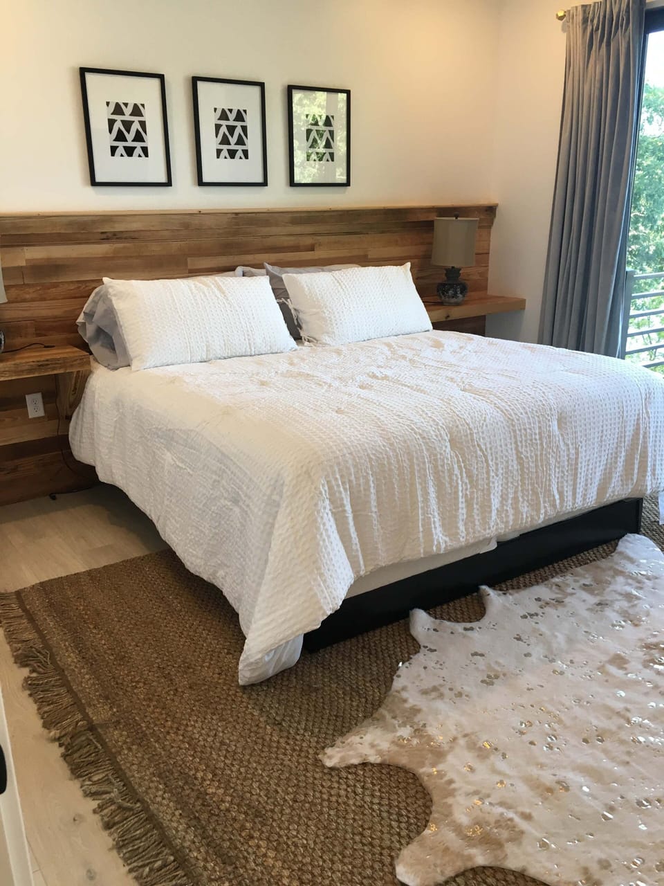 Relax in this peaceful bedroom featuring crisp bedding, warm wood furnishings, tasteful artwork, and a large window offering scenic views and plenty of natural light.