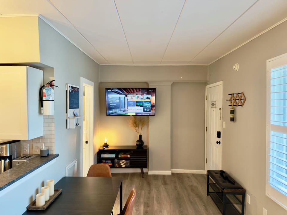 Open-concept living space featuring a modern kitchen, dining table, flat-screen TV and entryway with large windows. Neutral tones and clean design create a bright, inviting atmosphere perfect for cooking, dining and relaxation.