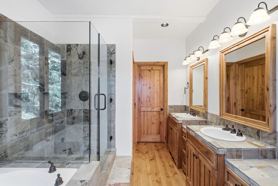 Modern bathroom with walk-in glass shower, double sinks, natural stone finishes, and large windows bringing in natural light